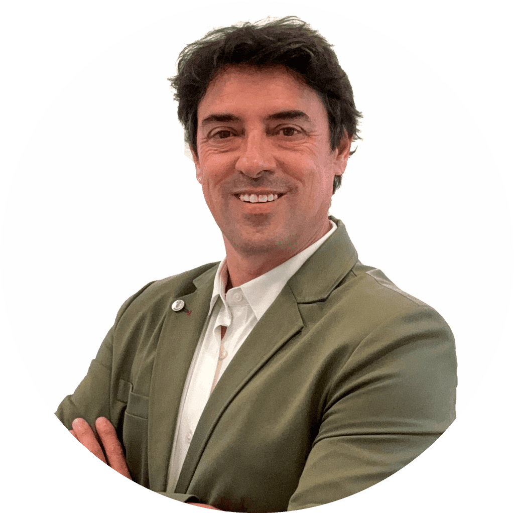 Un hombre vestido con una chaqueta verde y una camisa blanca sonríe con los brazos cruzados sobre un fondo blanco.