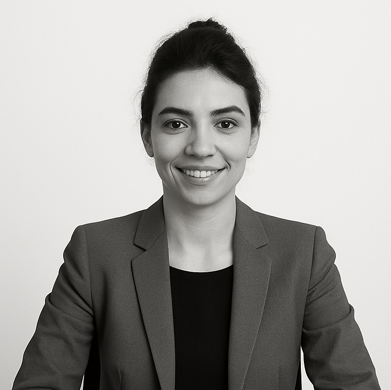 Una mujer vestida con americana está sentada en una mesa, mirando a la cámara y sonriendo, con las manos cruzadas sobre la superficie que tiene delante. El fondo es liso y de color claro.