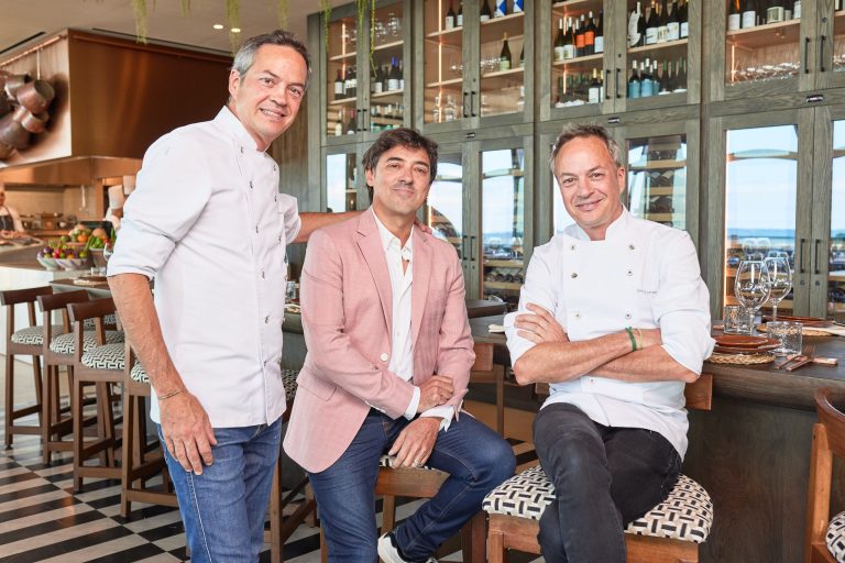 Tres hombres posan en un elegante restaurante, dos con bata de cocinero y uno con americana rosa, sentados y de pie frente a una barra con vitrinas llenas de botellas.